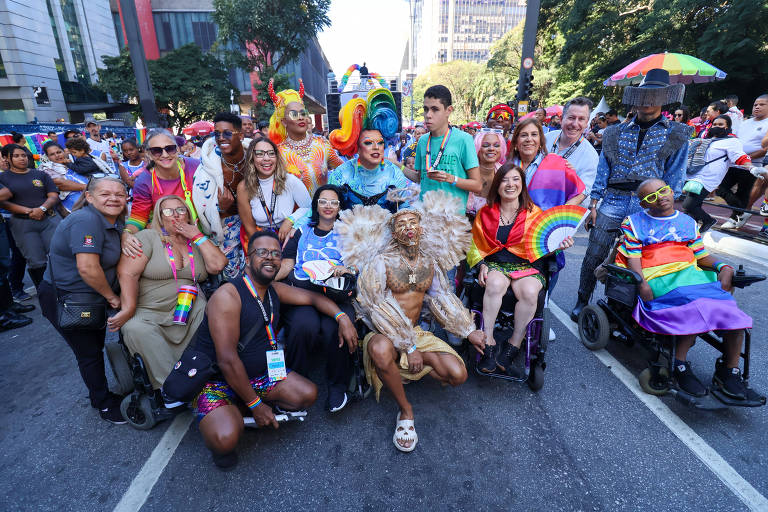 Um grupo diversificado de pessoas posa sorrindo durante parada do orgulho LGBTQ+, exibindo roupas coloridas e acessórios vibrantes. Eles estão cercados por bandeiras do arco-íris