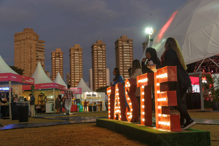 A imagem captura a atmosfera vibrante de um festival ao ar livre durante a noite, com pessoas sentadas em letras grandes iluminadas que formam a palavra "TASTE". Barracas coloridas e uma grande estrutura de tenda podem ser vistas ao fundo, enquanto arranha-céus 