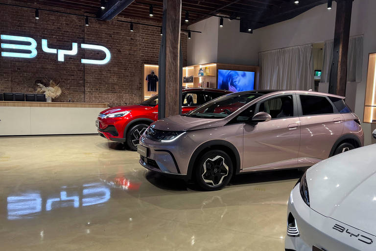 A imagem mostra um showroom iluminado com o logotipo "BYD" em destaque, exibindo carros elétricos modernos com design elegante. O piso brilhante reflete as luzes e os logotipos.