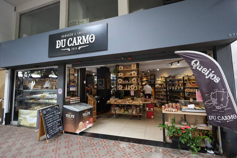 Fachada da loja Empório Du Carmo com sua entrada aberta. A imagem exibe uma variedade de queijos e embutidos, enquanto um quadro-negro do lado de fora anuncia ofertas especiais