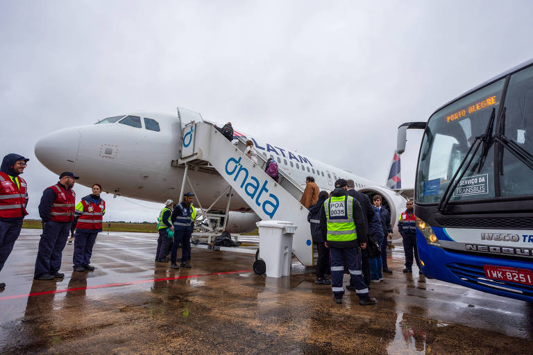 Na imagem vemos passageiros subindo uma escada de embarque de uma aeronave. Na direita, vemos alguns passageiros saindo de um ônibus estacionado.