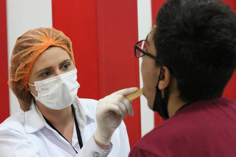 À esquerda, dentista usando touca laranja, máscara cirúrgica branca e luvas abre boca de paciente com instrumento. 