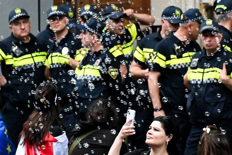 Um grupo de policiais uniformizados está cercado por uma nuvem de bolhas de sabão, enquanto uma mulher, em primeiro plano, tira uma selfie com a cena inusitada ao fundo. A expressão séria dos policiais contrasta com a leveza e alegria que as bolhas geralmente representam, criando uma cena curiosa e fotogênica.