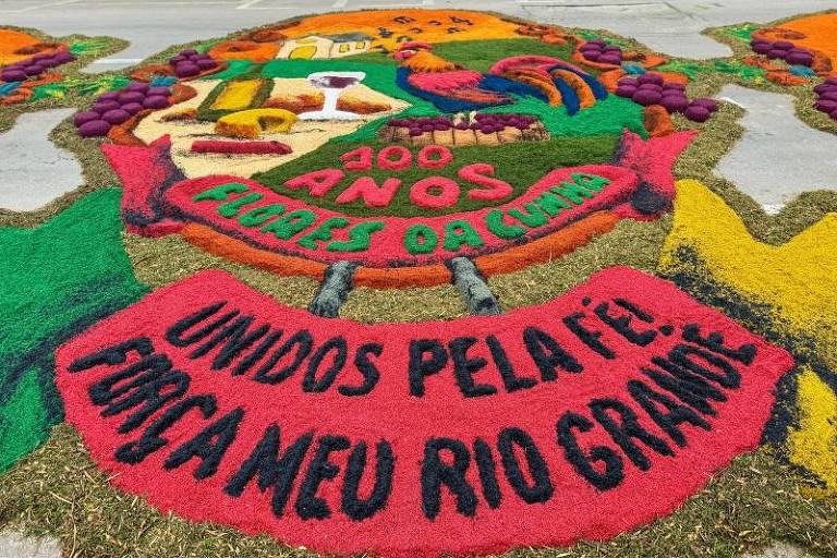 .Na foto, um tapete de serragem colorido em verde, vermelho e amarelo diz as frases "100 anos Flores da Cunha" e "Unidos pela fé! Força meu Rio Grande"