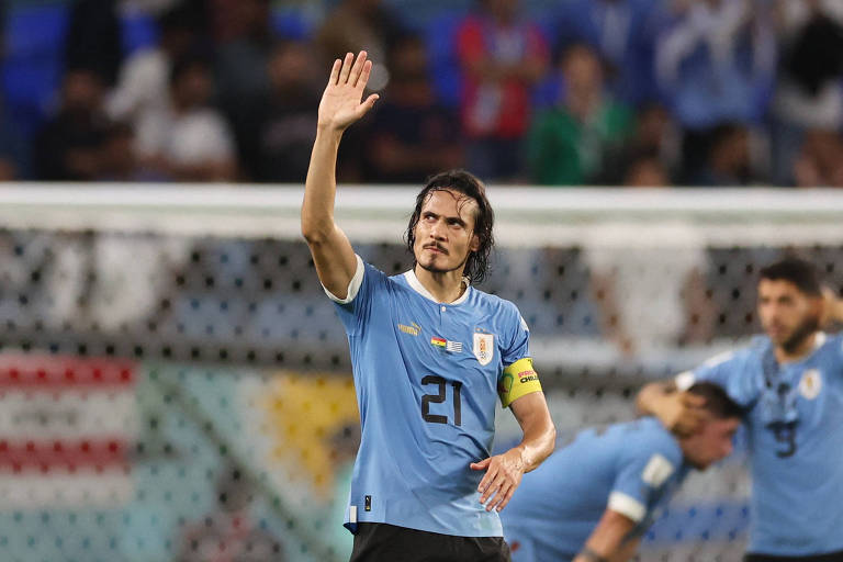 Homem com cabelos castanhos compridos e uniforme de futebol, com camisa azul e shorts preto, acena em direção à torcida. Ele tem um semblante sério. Atrás dele, é possível ver, com menos foco, dois outros jogadores se abraçando em frente ao gol