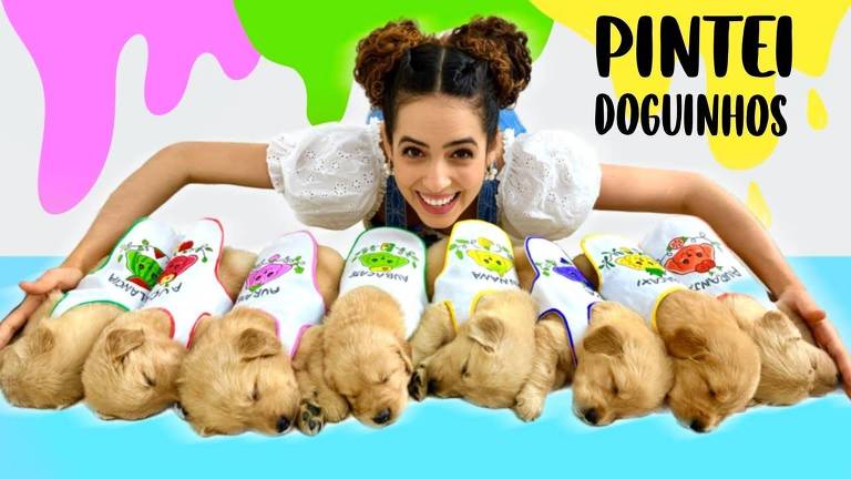 YouTuber Paula Stephânia sorridente com duas marias no cabelo está deitada atrás de uma fileira de filhotes de cachorro adormecidos, cada um vestindo uma camiseta branca com desenhos coloridos de frutas e legumes. O fundo é decorado com manchas coloridas.