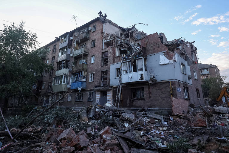 A imagem mostra um prédio residencial gravemente danificado, com várias seções da estrutura desmoronadas e escombros espalhados pelo chão. O céu azul ao fundo contrasta com a cena de devastação, sugerindo um momento de calma após um evento catastrófico.