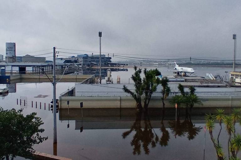 Na foto, prédios do aeroporto de Porto Alegre estão rodeados de água. Ao fundo, um avião pousado.
