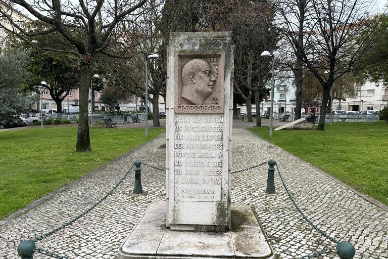 Um monumento com um relevo de perfil masculino ergue-se em um parque tranquilo, cercado por uma corrente verde e postes de metal. Árvores desfolhadas e um banco de parque ao fundo evocam uma atmosfera serena de um dia nublado.