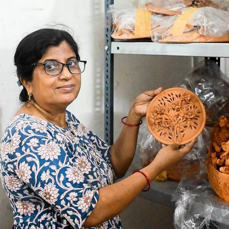 Uma mulher com óculos e uma blusa estampada azul e branca está de pé ao lado de uma prateleira de despensa, segurando uma peça de cerâmica redonda.