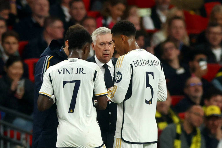 Homem de cabelos brancos e terno conversa com dois jogadores uniformizados na beira do campo em um estádio de futebol. Os jogadores estão de costas para a foto, vestem uniformes brancos e é possível ler "Vini Jr. 7" e "Bellingham 5" em suas camisas