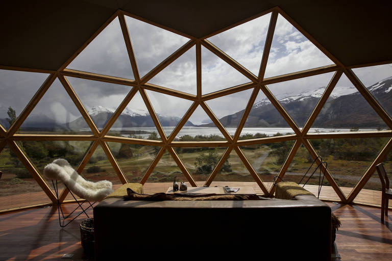 Imagem mostra acomodação, com um sofá, uma cadeira e uma mesa de centro, dispostos em espaço em forma de iglu, com estrutura de madeira e vidro com vista para natureza. Lago, montanhas e campo são vistos ao longe.