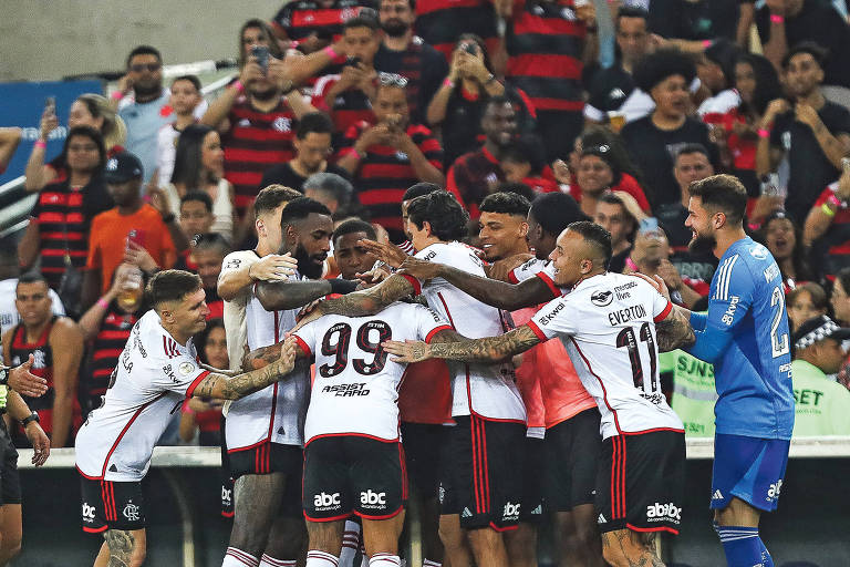 Jogadores com uniforme de camisa branca e shorts preto fazem abraço coletivo no gramado em comemoração a um dos gols; há também um goleiro vestido de azul; ao fundo, é possível ver a torcida na arquibancada vestida com camisas de cor predominantemente preta e vermelha