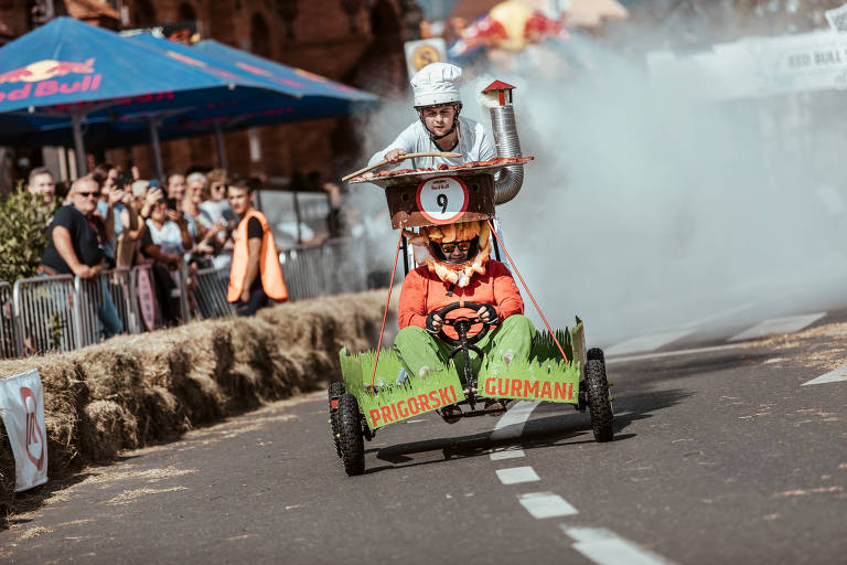 Um participante vestido com capacete e óculos de proteção pilota um carrinho de rolimã decorado com temas de culinárias, criando uma nuvem de fumaça ao fundo, enquanto espectadores observam a cena