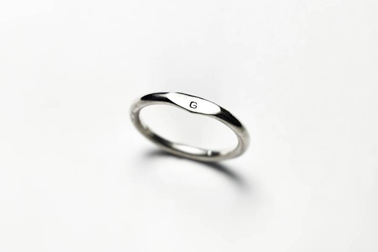 Um anel de prata, com letra "G" em destaque, em um fundo branco uniforme