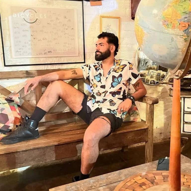 Um homem com barba e cabelo curto, vestindo uma camisa estampada e shorts, está sentado descontraidamente em um banco de madeira. Ele parece estar em um ambiente com decoração vintage, incluindo um globo terrestre ao seu lado