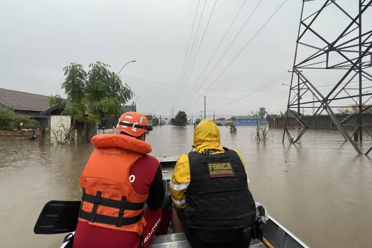 Dois socorristas em um bote navegam por uma área residencial inundada, com águas que submergem parcialmente as casas e a vegetação local. O céu nublado e a água se estende até onde a vista alcança. Os socorristas estão equipados com coletes salva-vidas e roupas de chuva