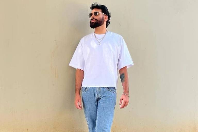 Um homem com barba e cabelo curto, vestindo uma camiseta branca simples, calça jeans azul clara e tênis brancos, está de pé contra uma parede de cor clara. Ele olha para cima com uma expressão relaxada