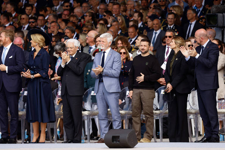 Zelenski, entre o presidente tcheco, Petr Pavel (de cinza claro) e sua mulher Olena, agradece aos aplausos após ser citado por Emmanuel Macron no evento do Dia D na praia de Omaha