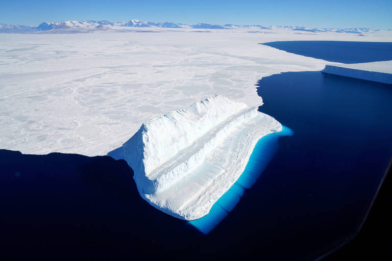 A imagem captura a beleza imponente de um iceberg, com sua massa branca contrastando com o azul profundo do oceano. A luz do sol ilumina a superfície gelada, revelando nuances de azul e branco, enquanto o horizonte distante mostra a vastidão da paisagem polar.