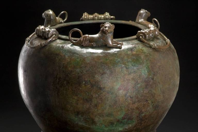 A imagem mostra um antigo vaso de bronze com uma pátina verde-azulada, indicando idade e oxidação. Quatro figuras de animais adornam a borda superior, servindo como pontos de fixação para o que parece ser uma alça quebrada ou ausente. O vaso repousa sobre um suporte que o eleva, destacando sua forma arredondada e as características decorativas que sugerem uma origem em uma antiga civilização.