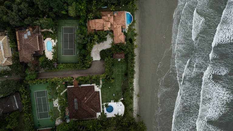 Imagem aérea mostra casas com piscinas e quadras de tênis perto da faixa de areia com o mar adiante