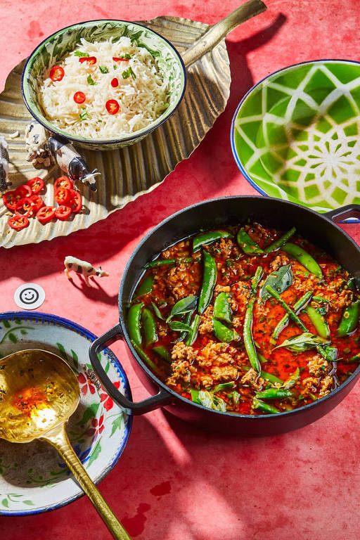 A foto mostra um pequeno caldeirão preto com um cozido de curry com carne moída vegana sobre uma base vermelha. Ao lado, uma porção de arroz com fatias de pimenta vermelha