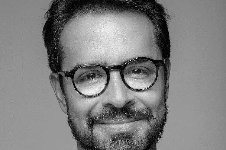 Homem com barba, cabelos lisos e escuros e óculos de armação grossa. O retrato é em preto e branco com fundo neutro