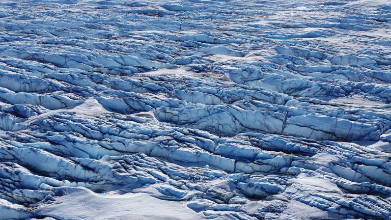 A imagem captura a vastidão de um terreno coberto de gelo, com a luz do sol refletindo sobre a superfície irregular e azulada, criando um mosaico de sombras e texturas que evocam a beleza e a severidade dos ambientes polares.