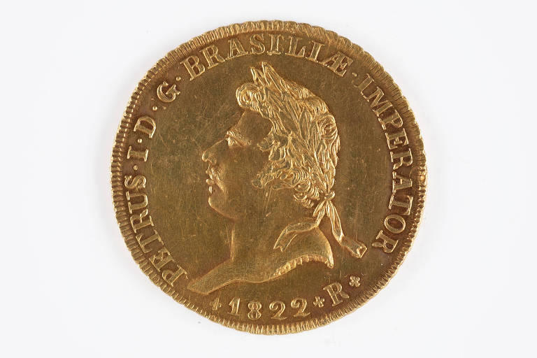 A imagem mostra uma moeda dourada antiga do Brasil, com o perfil de um homem olhando para a esquerda, cercado por inscrições que indicam sua origem e valor. A moeda é datada de 1822 e possui marcas que sugerem sua circulação e uso ao longo do tempo.
