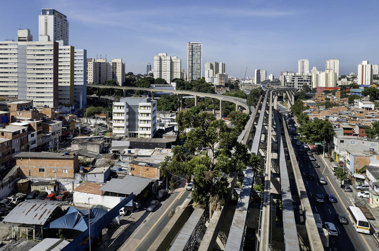 A imagem mostra trilhos suspensos, atravessados por uma árvores, que lembram um viaduto de concreto. Ele se estende por cima de uma área com habitações precárias, contrastando com prédios modernos ao fundo sob um céu claro.