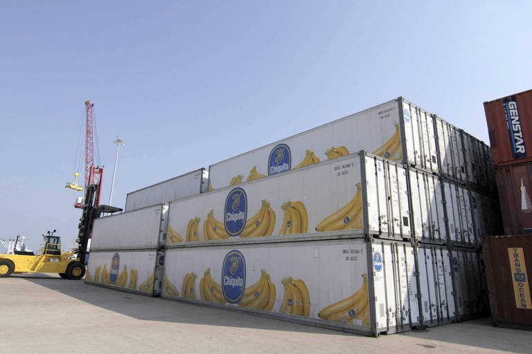 Três contêineres brancos com desenhos de bananas aparecem empilhados em um porto. Máquinas aparecem trabalhando ao fundo.