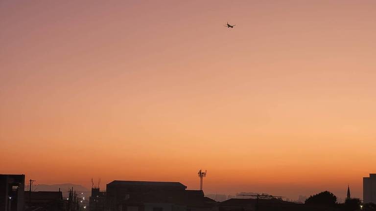 O céu exibe um degradê de cores quentes, variando do laranja ao azul, enquanto o sol se põe atrás de silhuetas urbanas. Um avião passa no céu vasto