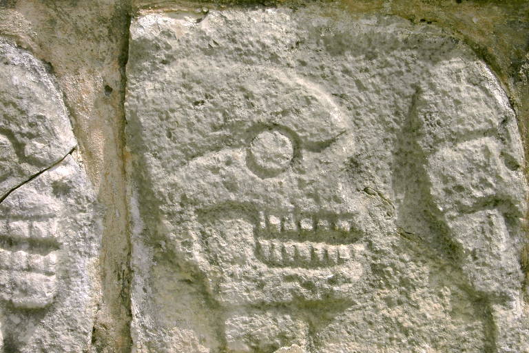 A imagem mostra um relevo em pedra com duas figuras esculpidas que parecem representar rostos estilizados ou máscaras. A textura desgastada e a aparência antiga sugerem que se trata de um artefato arqueológico, possivelmente de uma civilização antiga.