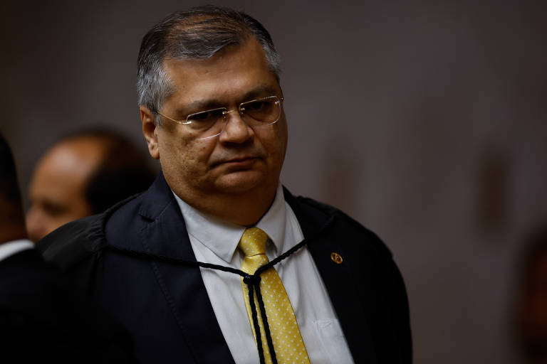 Imagem mostra Flávio Dino, um homem com cabelo curto e grisalho, usando óculos, está vestido com um terno escuro e uma gravata amarela. Ele parece estar em um ambiente formal, com uma expressão séria. Ao fundo, é possível ver outras pessoas, mas não estão claramente visíveis.