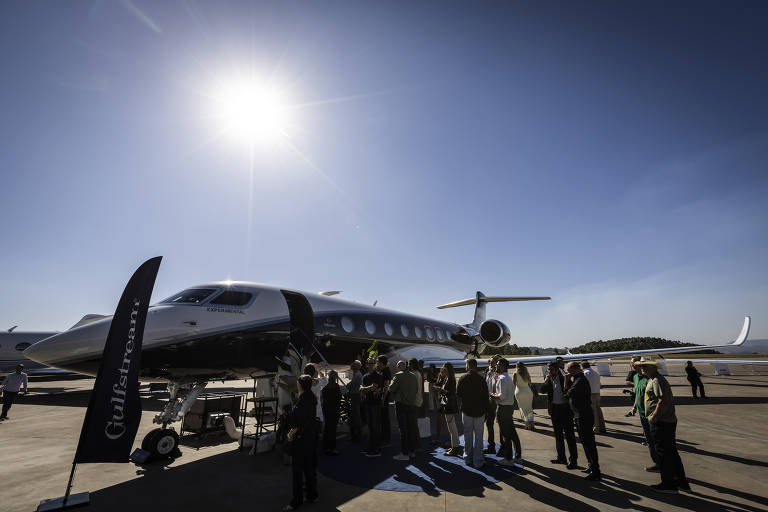 Pessoas rodeiam jato executivo em pista de aeroporto. Sol aparece em céu sem nuvens.