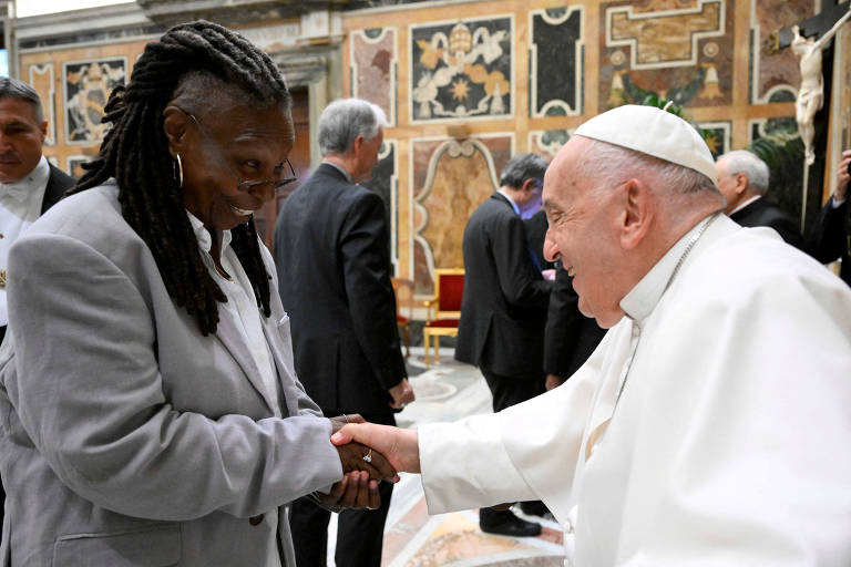 Em um ambiente adornado com afrescos e mosaicos, uma mulher sorridente com tranças cumprimenta calorosamente o Papa, que retribui com um sorriso igualmente afetuoso. O momento de cordialidade entre os dois destaca-se em meio a outros indivíduos ao fundo, sugerindo um encontro significativo em um contexto formal.