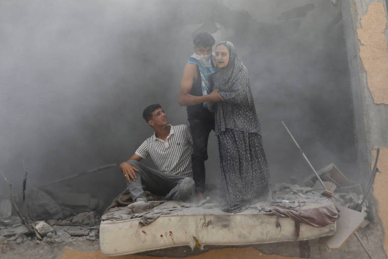 Três pessoas, um homem sentado, uma mulher de pé e uma criança no colo, cercados por poeira e destroços, parecem desolados após um evento destrutivo. Eles estão sobre o que parece ser um colchão sujo, com a mulher consolando a criança e o homem olhando para o lado, possivelmente em busca de ajuda ou observando a extensão dos danos.