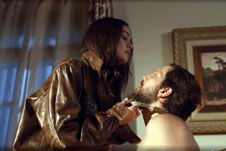 A imagem captura um momento de intimidade entre um homem e uma mulher; ela, vestindo uma jaqueta de couro, segura no queixo dele, sem camisa