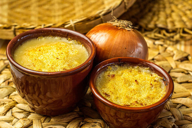 Duas cumbucas pequenas com sopa de cebola gratinada. Entre elas, há uma cebola inteira.