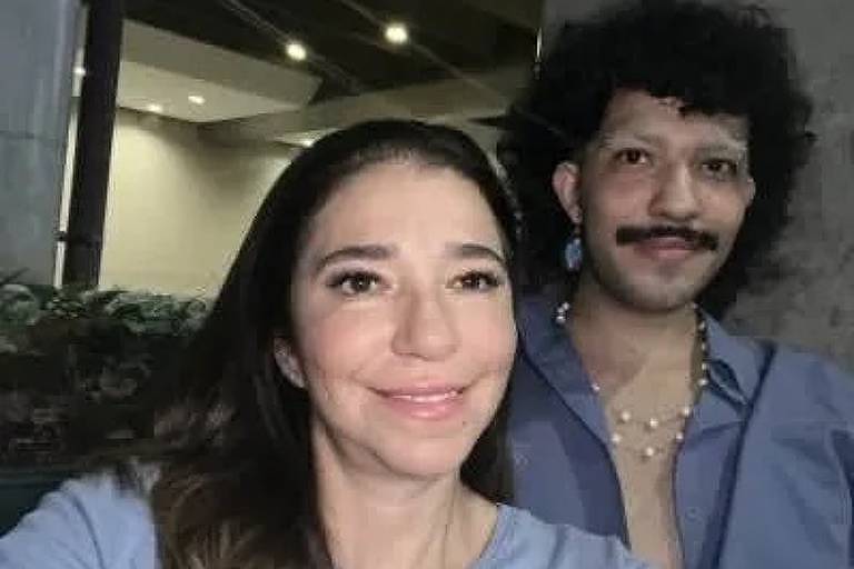 Mulher e homem posa para uma selfie aparentemente noturna, com a mulher à frente sorrindo para a câmera e o homem ao fundo exibindo um bigode e cabelos encaracolados, ambos parecendo felizes e descontraídos em um ambiente externo com iluminação suave.