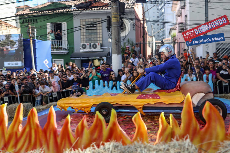 Um participante vestido com um macacão azul e capacete de segurança pilota um carrinho de corrida artesanal que imita uma fatia de pizza gigante. Ele parece estar se divertindo enquanto manobra o veículo através de um percurso decorado com chamas falsas, sob o olhar atento de uma multidão que o cerca.