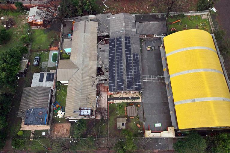 A imagem captura uma visão aérea de uma área residencial após uma tempestade, mostrando uma casa com o telhado parcialmente destruído e destroços espalhados pelo quintal. Ao lado, um grande edifício amarelo com um telhado intacto contrasta com a cena de destruição.