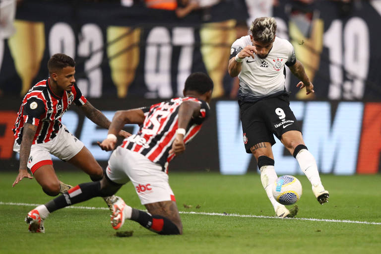 Três jogadores de futebol em um momento intenso de jogo, com um atleta do time de uniforme preto e branco controlando a bola enquanto dois adversários do time listrado tentam interceptar a jogada.
