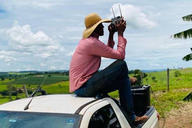 Um homem senta-se no teto de um carro, sob um céu parcialmente nublado, cercado por uma paisagem verdejante.