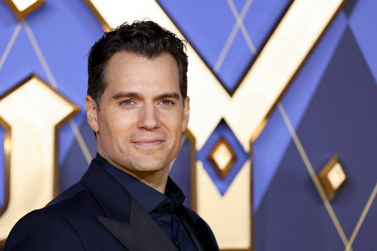 A foto mostra o ator Henry Cavill, um homem branco de cabelo castanho escuro vestindo um smoking preto com um leve sorriso. Atrás dele, há um fundo gráfico de uma première de um filme