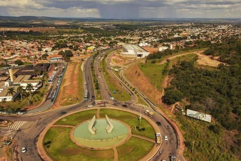 A imagem mostra uma vista aérea de um cruzamento onde uma linha ferroviária atravessa uma rodovia. No centro, um canteiro circular com um monumento é o ponto focal, enquanto as estradas se estendem para áreas urbanas ao fundo.