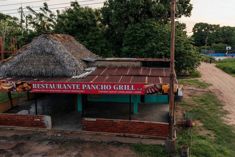 Um pequeno restaurante com um telhado de palha e uma fachada colorida, o 'Restaurante Pancho Grill', oferece um vislumbre da culinária local ao lado de uma estrada de terra, evocando uma atmosfera rústica e acolhedora em um ambiente tropical.
