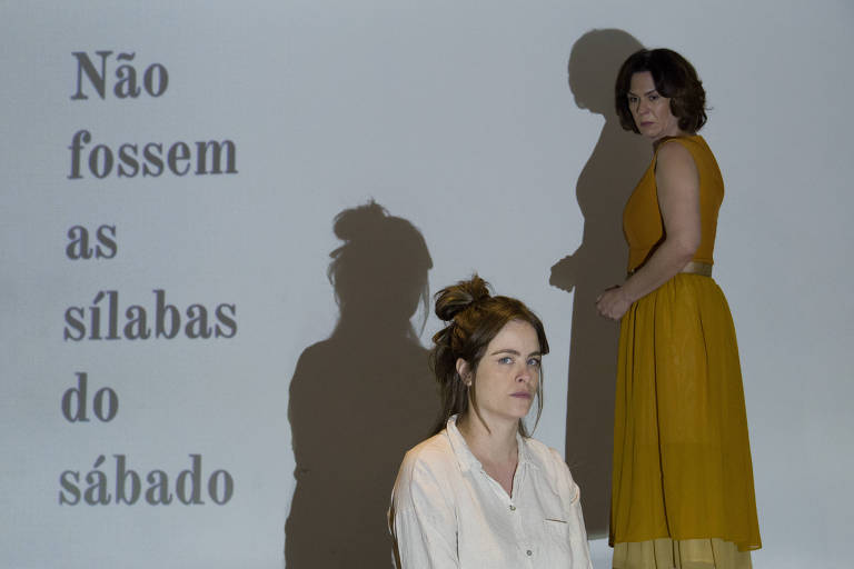 Duas mulheres estão em frente a uma parede onde a frase "Não Fossem as Sílabas do Sábado" em é projetada. A mulher à esquerda usa uma camisa branca e olha para o lado, enquanto a mulher à direita, vestida com uma saia amarela e uma blusa sem mangas, olha diretamente para a câmera com uma expressão séria