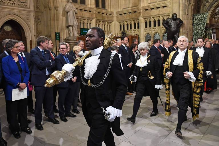 Ugbana, um homem negro vestido com trajes cerimoniais, incluindo uma corrente e um bastão, lidera um grupo de pessoas em trajes formais por um corredor adornado com arquitetura gótica. Os espectadores, alinhados ao longo do corredor, observam a procissão com expressões de respeito e interesse.